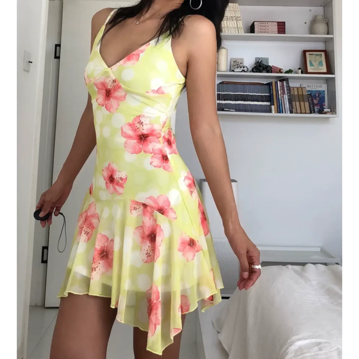 Angled view of a woman wearing a yellow mesh mini dress with pink hibiscus flowers and white polka dots, showing the fitted bodice and asymmetrical ruffled hem.