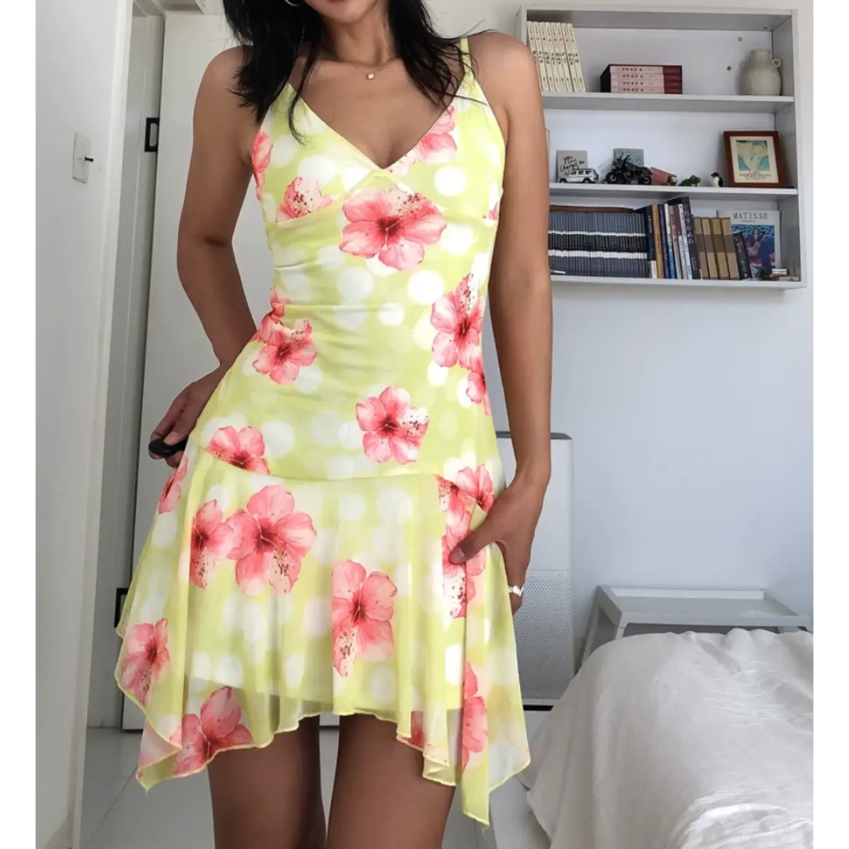 Woman wearing a form-fitting yellow mesh mini dress with pink hibiscus flowers and white polka dots, featuring spaghetti straps, a V-neckline, and an asymmetrical ruffled hem.