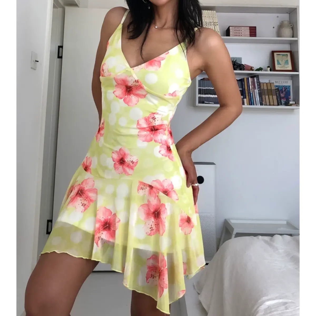 Woman wearing a yellow mesh mini dress with pink hibiscus flowers and white polka dots, posed with one hand on her hip to highlight the dress's silhouette.

