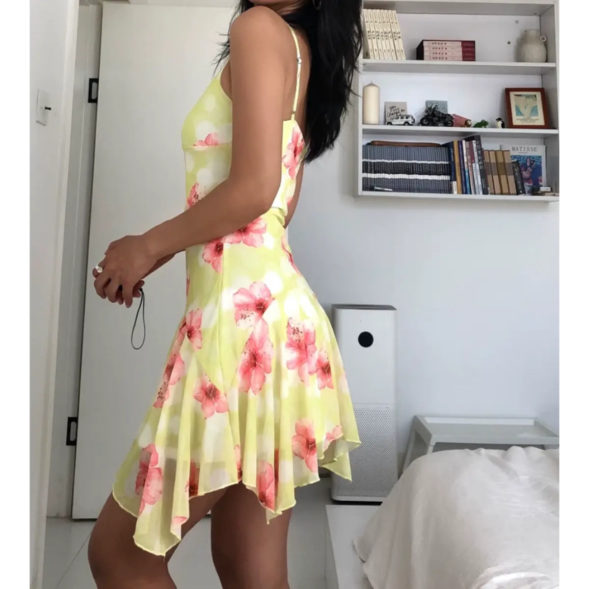 Three-quarter back view of a woman wearing a yellow mesh mini dress with pink hibiscus flowers and white polka dots, showing the open back and asymmetrical hem.
