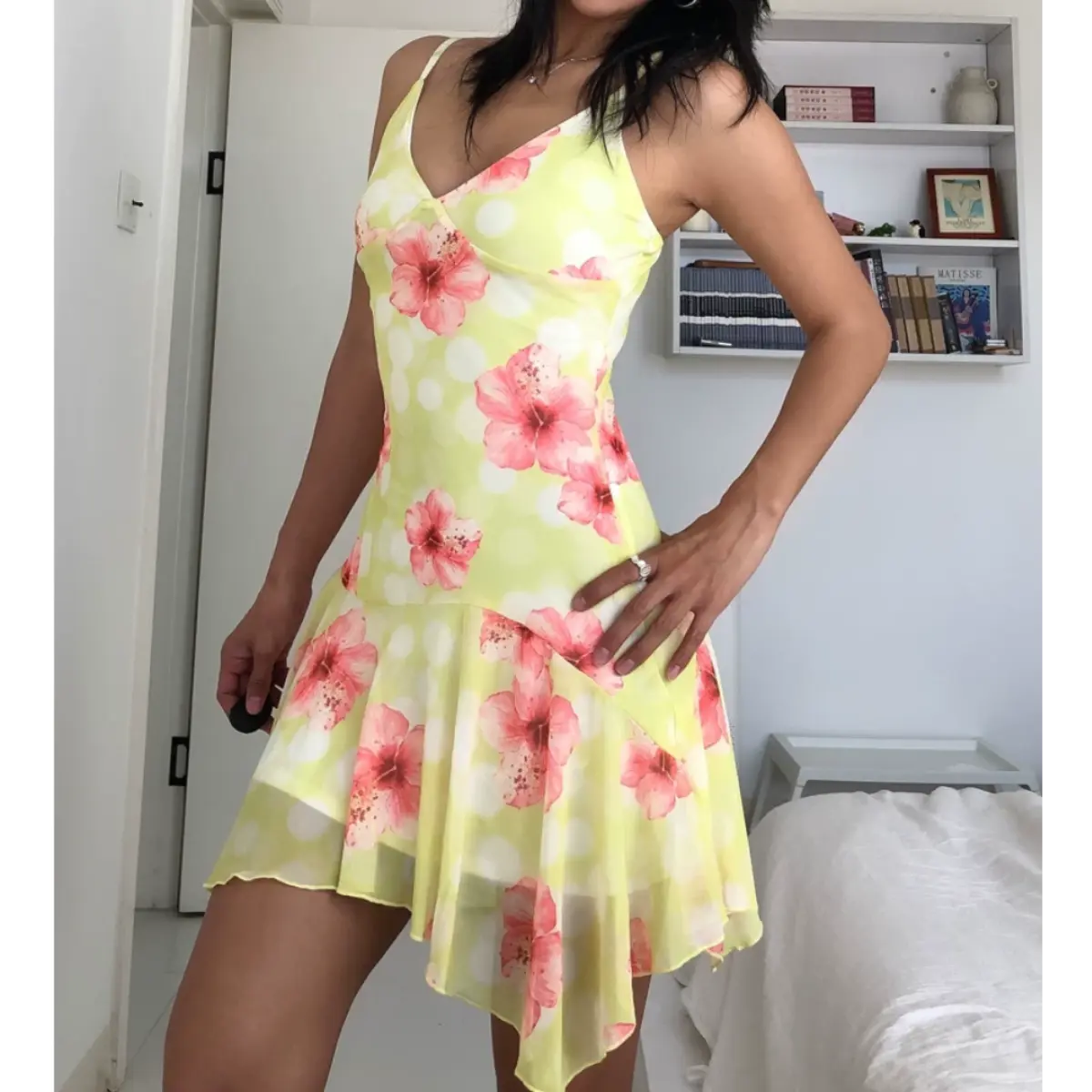 Three-quarter view of a woman wearing a yellow mesh mini dress with pink hibiscus flowers and white polka dots, showing the side profile and asymmetrical hem.