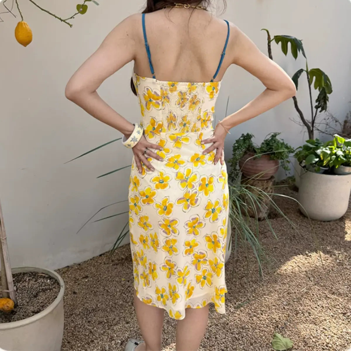 Back view of a woman wearing a cream midi slip dress with a yellow watercolor floral print, blue spaghetti straps, and a smocked back panel, styled outdoors.
