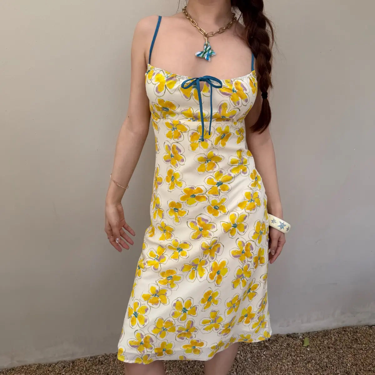  Front view of a woman in a cream midi slip dress with a yellow watercolor floral print, featuring blue spaghetti straps and a matching blue bow at the bust.