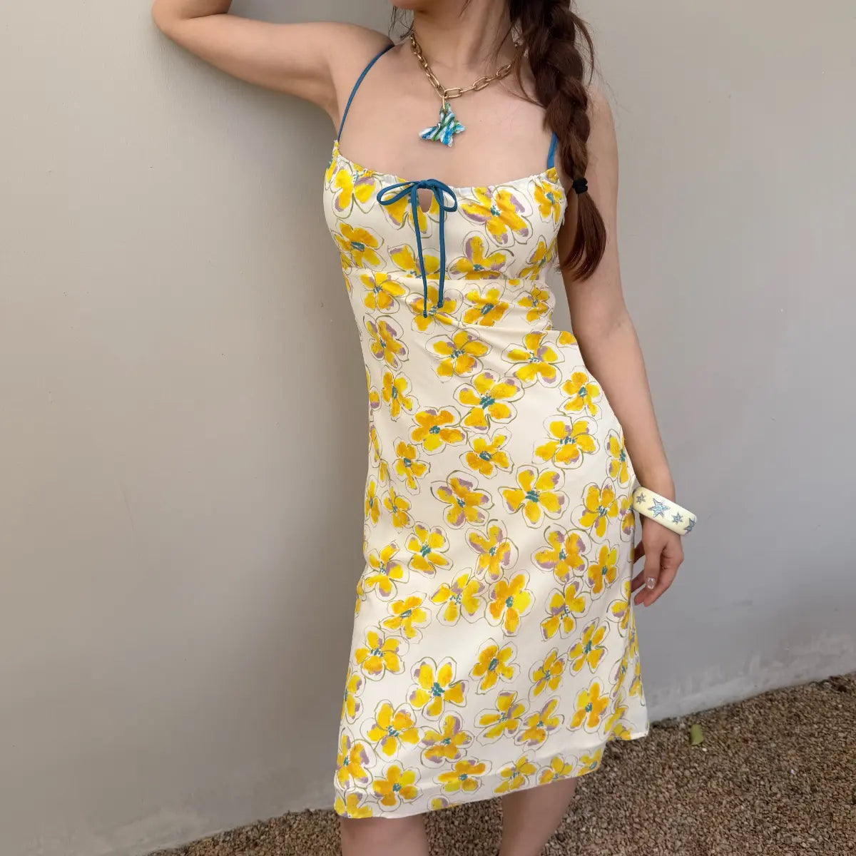 Woman in a cream midi slip dress with a yellow watercolor floral print, styled with a braid and accessories, leaning against a wall.