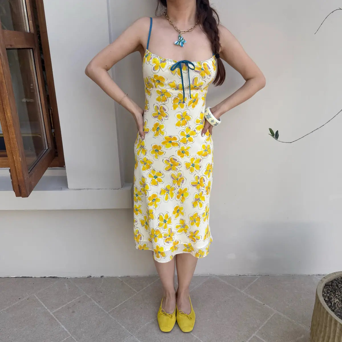 Full view of a woman in a cream midi slip dress with a yellow watercolor floral print, paired with yellow shoes, standing by a window.