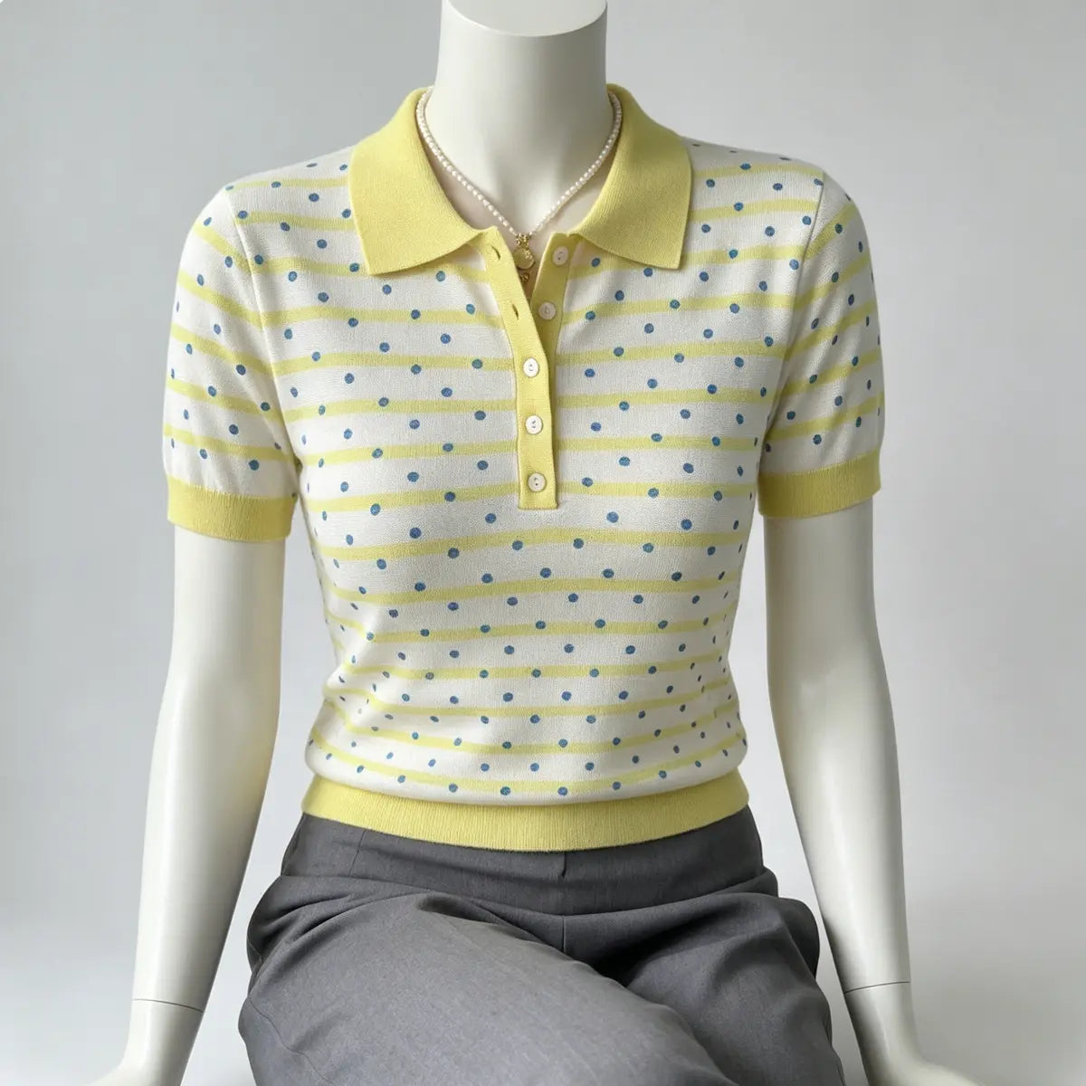 Mannequin wearing a yellow and white polka dot knit polo shirt, featuring ribbed yellow collar, cuffs, and hem, paired with grey trousers.